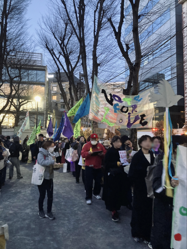 ウィメンズマーチ東京2026の出発前の様子。思い思いのプラカードや、「VOTE FOR LOVE」と書かれた横断幕など、色とりどりの大きな旗を持った人が並ぶ。